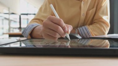 Bir insan beyaz bir kalemle bir tablete yazı yazıyor. Tablet ahşap bir masanın üzerinde.