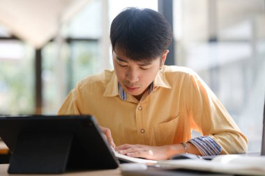 Sınavlara hazırlanmak için çevrimiçi öğrenim gören, notlar alan ve çalışma materyallerini kitaplarla ve dijital cihazlarla inceleyen bir öğrenci.