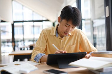 Sınavlara hazırlanmak için çevrimiçi öğrenim gören, notlar alan ve çalışma materyallerini kitaplarla ve dijital cihazlarla inceleyen bir öğrenci.