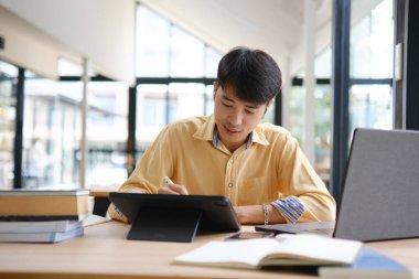 Sınavlara hazırlanmak için çevrimiçi öğrenim gören, notlar alan ve çalışma materyallerini kitaplarla ve dijital cihazlarla inceleyen bir öğrenci.