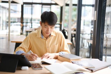 Sınavlara hazırlanmak için çevrimiçi öğrenim gören, notlar alan ve çalışma materyallerini kitaplarla ve dijital cihazlarla inceleyen bir öğrenci.