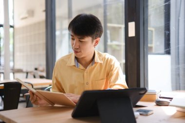 Sınavlara hazırlanmak için çevrimiçi öğrenim gören, notlar alan ve çalışma materyallerini kitaplarla ve dijital cihazlarla inceleyen bir öğrenci.