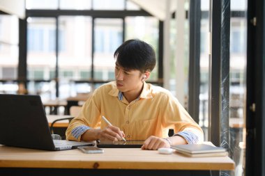 Sınavlara hazırlanmak için çevrimiçi öğrenim gören, notlar alan ve çalışma materyallerini kitaplarla ve dijital cihazlarla inceleyen bir öğrenci.