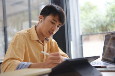 Sınavlara hazırlanmak için çevrimiçi öğrenim gören, notlar alan ve çalışma materyallerini kitaplarla ve dijital cihazlarla inceleyen bir öğrenci.