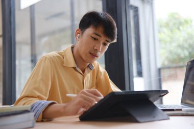 Sınavlara hazırlanmak için çevrimiçi öğrenim gören, notlar alan ve çalışma materyallerini kitaplarla ve dijital cihazlarla inceleyen bir öğrenci.