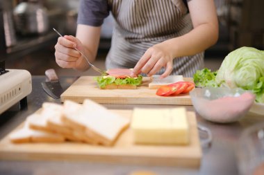 Profesyonel bir mutfakta bir şef, gurme sandviçi hazırlarken malzemeleri katmanlamaya konsantre olur. Ön planda renkli sebzeler ve ekmekler var, canlı bir görsel derinlik katıyor..