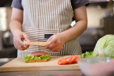 Profesyonel bir mutfakta bir şef, gurme sandviçi hazırlarken malzemeleri katmanlamaya konsantre olur. Ön planda renkli sebzeler ve ekmekler var, canlı bir görsel derinlik katıyor..
