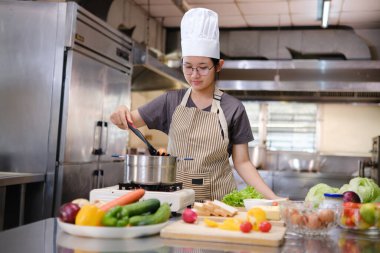 Odaklanmış genç bir aşçı, profesyonel bir mutfağın gaz ocağında yumurta karıştırıyor ve tezgahın üzerinde taze ürünler ve sandviç malzemeleri düzgünce düzenleniyor..