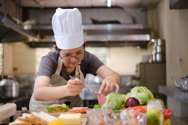 Profesyonel bir mutfakta bir şef, gurme sandviçi hazırlarken malzemeleri katmanlamaya konsantre olur. Ön planda renkli sebzeler ve ekmekler var, canlı bir görsel derinlik katıyor..