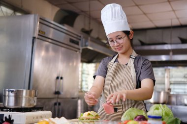 Şef, tezgahtaki taze sebzelerle çevrili sandviçin üzerine dikkatlice bir dilim jambon koyar..