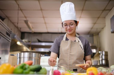 Profesyonel bir mutfakta bir şef, gurme sandviçi hazırlarken malzemeleri katmanlamaya konsantre olur. Ön planda renkli sebzeler ve ekmekler var, canlı bir görsel derinlik katıyor..