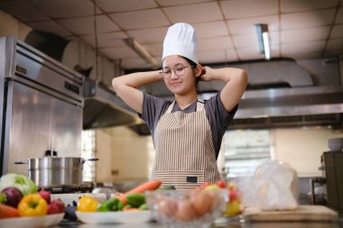 Profesyonel bir aşçı, aşçı şapkasını düzelterek mutfak işlerine hazırlanır. Yemek pişirme sürecine başlamadan önce yeni malzemelerle çevrili ticari bir mutfakta durur..