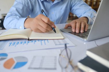 Profesyonel işadamı, ofis masasındaki mali tabloları incelerken dizüstü bilgisayarda not alıp daktilo kullanıyor.