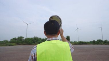 Kask ve güvenlik yeleği giyen mühendis, yenilenebilir enerji alanındaki rüzgar değirmenlerini gözlemliyor, operasyonel kontroller yapıyor ve temiz enerji performansı sağlıyor. Yüksek kalite 4k görüntü