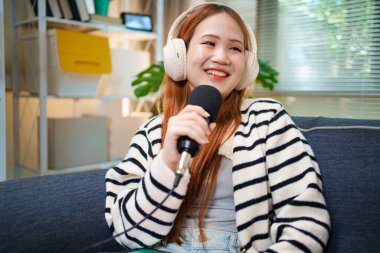 Evde podcast yaparken gülümseyen genç bir kadın. Kapalı alanda mikrofon ve kulaklık kullanıyor..