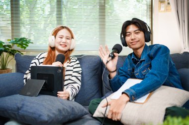 Genç bir adam ve kadın evlerinde mikrofon ve kulaklıklarla podcast yaparken kameraya el sallıyor, rahat bir oturma odasında kanepede oturuyor..