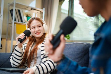 Genç adam ve kadın koltukta otururken mikrofon ve kulaklık kullanarak podcast yapıyorlar..