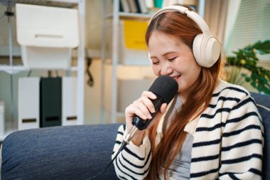 Evde podcast yaparken gülümseyen genç bir kadın. Kapalı alanda mikrofon ve kulaklık kullanıyor..