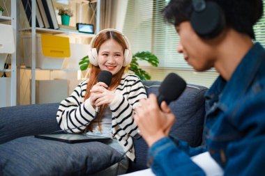 Genç adam ve kadın koltukta otururken mikrofon ve kulaklık kullanarak podcast yapıyorlar..