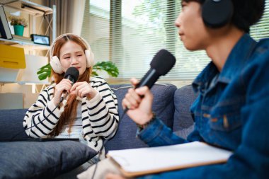 Genç adam ve kadın koltukta otururken mikrofon ve kulaklık kullanarak podcast yapıyorlar..