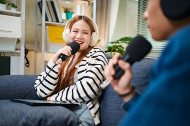 Genç adam ve kadın koltukta otururken mikrofon ve kulaklık kullanarak podcast yapıyorlar..