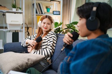 Genç adam ve kadın koltukta otururken mikrofon ve kulaklık kullanarak podcast yapıyorlar..