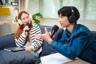 Genç adam ve kadın koltukta otururken mikrofon ve kulaklık kullanarak podcast yapıyorlar..