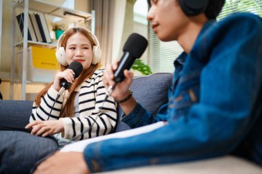 Genç adam ve kadın koltukta otururken mikrofon ve kulaklık kullanarak podcast yapıyorlar..