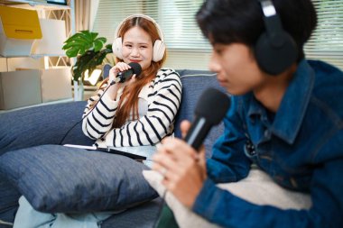 Genç adam ve kadın koltukta otururken mikrofon ve kulaklık kullanarak podcast yapıyorlar..