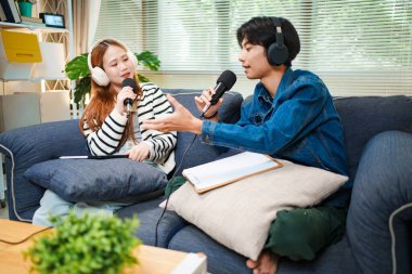 Genç adam ve kadın koltukta otururken mikrofon ve kulaklık kullanarak podcast yapıyorlar..