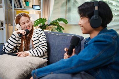 Genç adam ve kadın koltukta otururken mikrofon ve kulaklık kullanarak podcast yapıyorlar..