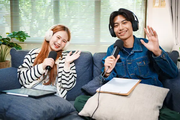 Genç bir adam ve kadın evlerinde mikrofon ve kulaklıklarla podcast yaparken kameraya el sallıyor, rahat bir oturma odasında kanepede oturuyor..