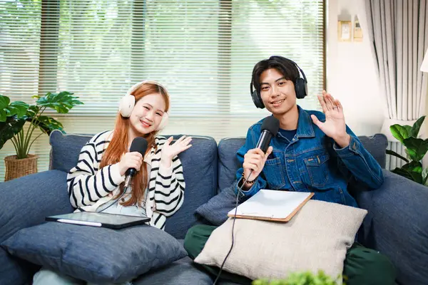 Genç bir adam ve kadın evlerinde mikrofon ve kulaklıklarla podcast yaparken kameraya el sallıyor, rahat bir oturma odasında kanepede oturuyor..