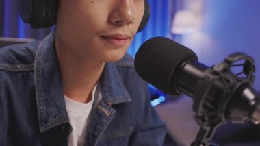 Genç bir Asyalı podcaster ya da sözü geçen kişi canlı yayın sırasında arka planda konuşurken, profesyonel mikrofonun aşırı yakın plan görüntüleri odaklı..