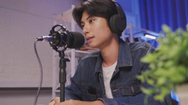 Yaratıcı bir ev stüdyosunda odaklanmış canlı yayın ya da podcast seansı sırasında kulaklık takan ve mikrofona konuşan Asyalı bir genç.