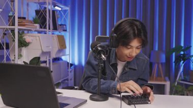 Kulaklık takan odaklanmış genç bir adam, yaratıcı mavi ışıklı ev stüdyosunda podcast kaydı için mikrofon ayarlıyor..