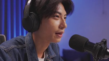 Genç Asyalı erkek etkili kulaklık takıyor, canlı yayın sırasında mikrofona ciddi şekilde konuşuyor veya modern bir ev stüdyosunda podcast yapıyor.