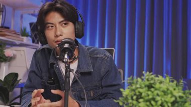 Genç Asyalı erkek içerik yaratıcısı kulaklık takıyor ve podcast veya canlı yayın sırasında profesyonel bir mikrofona dikkatle konuşuyor..