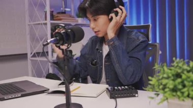 Kendine güvenen genç Asyalı adam modern bir ev stüdyosunda canlı yayın yapıyor kulaklık takıyor, mikrofon ayarlıyor ve ses mikserini ayarlıyor ve dizüstü bilgisayarında kayıt yapmaya başlıyor.