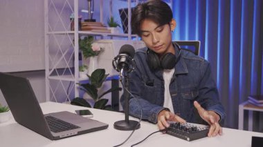 Asyalı içerik oluşturucu, özel ev stüdyosunda mikrofon, dizüstü bilgisayar ve ses karıştırıcı ile podcast düzeneği hazırlıyor.