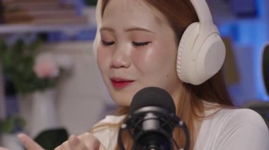 Neşeli genç Asyalı kadın radyo sunucusu ya da kulaklıklı podcaster flamalı profesyonel mikrofonla konuşan ve ev stüdyosunda yayın yapan