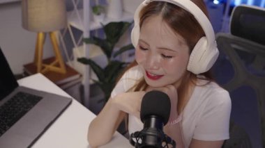 Kadın podcast sunucusu, mikrofona eğilirken konuşuyor. Modern stüdyo masasında yüksek açılı, sıcak ışıklandırmayla çekilmiş.