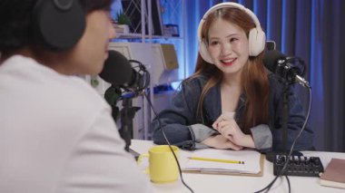 Bir podcast sunucusu, modern bir ev stüdyosundaki bir kayıt sırasında bir misafirle konuşurken dikkatle gülümser ve dinler..