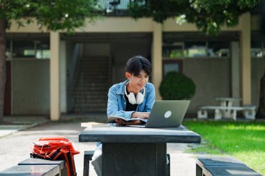Kampüste okuyan genç bir öğrenci. Erkek üniversite öğrencisi kampüs çimlerinde dizüstü bilgisayarda ödev yapıyor..