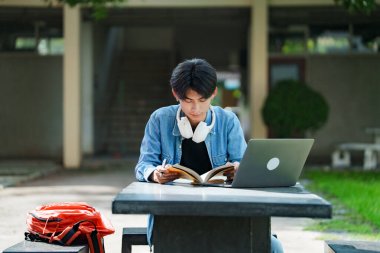 Kampüste okuyan genç bir öğrenci. Erkek üniversite öğrencisi kampüs çimlerinde dizüstü bilgisayarda ödev yapıyor..