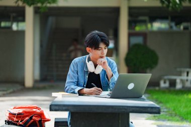 Kampüste okuyan genç bir öğrenci. Erkek üniversite öğrencisi kampüs çimlerinde dizüstü bilgisayarda ödev yapıyor..