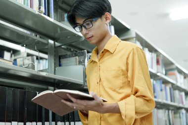Eğitim, öğrenim ve araştırma için bir kütüphanede kitap okuyan genç bir Asyalı. Okul, üniversite ya da üniversite kampüsünde. Dil ve felsefe öğrenmek için kitap rafında odaklan, kitap ve öğrenci