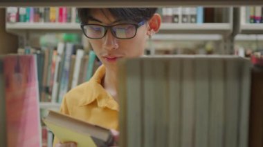 Kolej kütüphanesinde yürüyen ve kitaptan ders kitaplarını alan orta boy erkek öğrenciyi belden yukarı çek. Erkek Asyalı öğrenci arka planda kitaba göz atıyor.