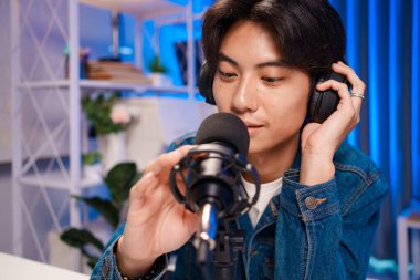 Genç adam evdeki stüdyoda dizüstü bilgisayar, kulaklık ve ses mikserini mavi ışık altında ayarlayarak podcast hazırlıyor..