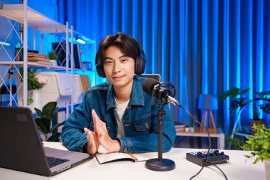 Bilgisayarında kulaklık ve mikroskopla podcast kaydeden bir adam. Parlak stüdyosundan canlı yayın yapan erkek podcaster